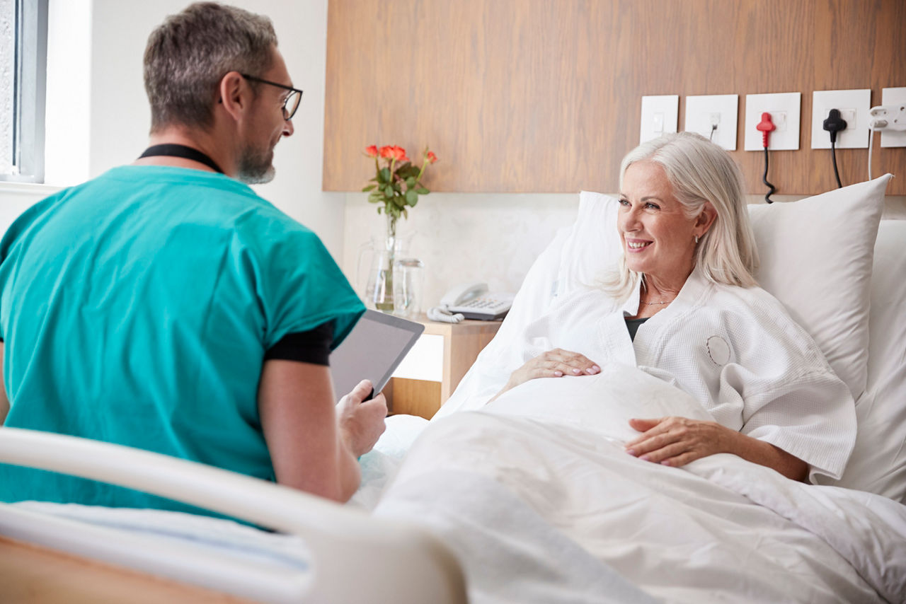 Arzt mit Tablet in der Hand macht Visite bei einer Patientin im Krankenzimmer an ihrem Bett