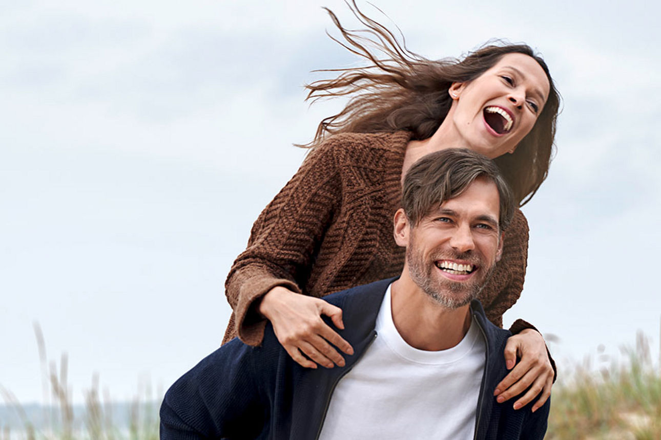 Ein lächelnder Mann trägt seine lächelnde Freundin auf dem Rücken an der Küste entlang.