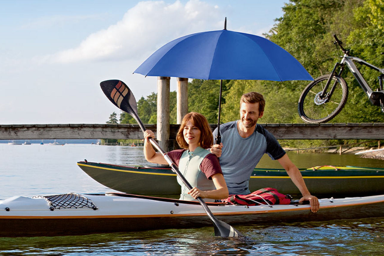 Frau und Mann in einem Kanu, mit Paddel und blauem Schirm an einer Anlegestelle