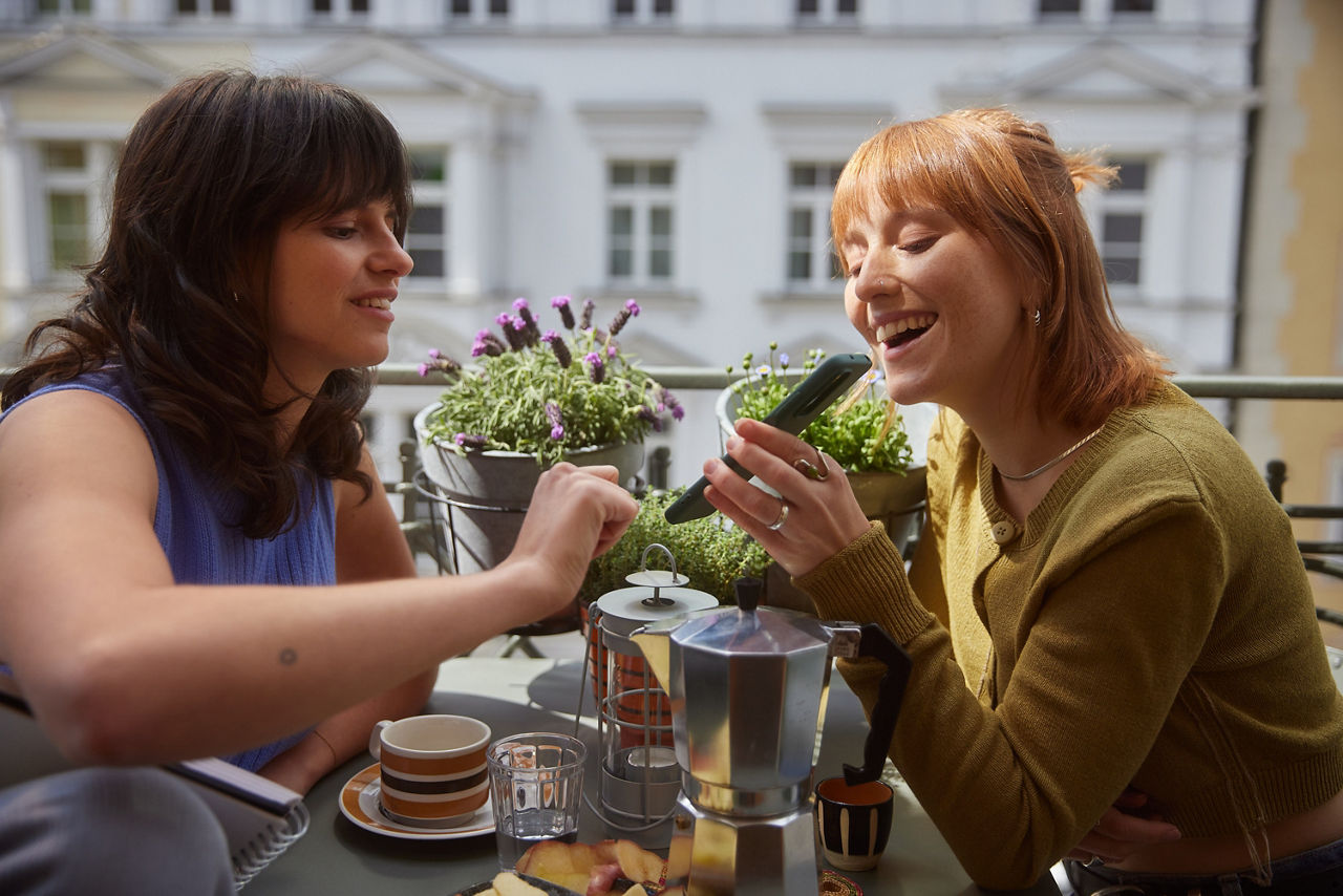 Zwei junge Frauen sitzen sich auf einem Balkon an einem mit Snacks und Kaffee gedecktem Tisch gegenüber. Die Rechte zeigt der Linken etwas auf ihrem Handy. 
