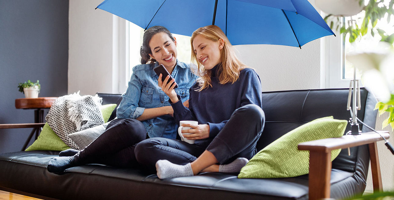 Zwei Frauen sitzen unter einem blauen Schirm auf dem Sofa und schauen lächelnd aufs Smartphone