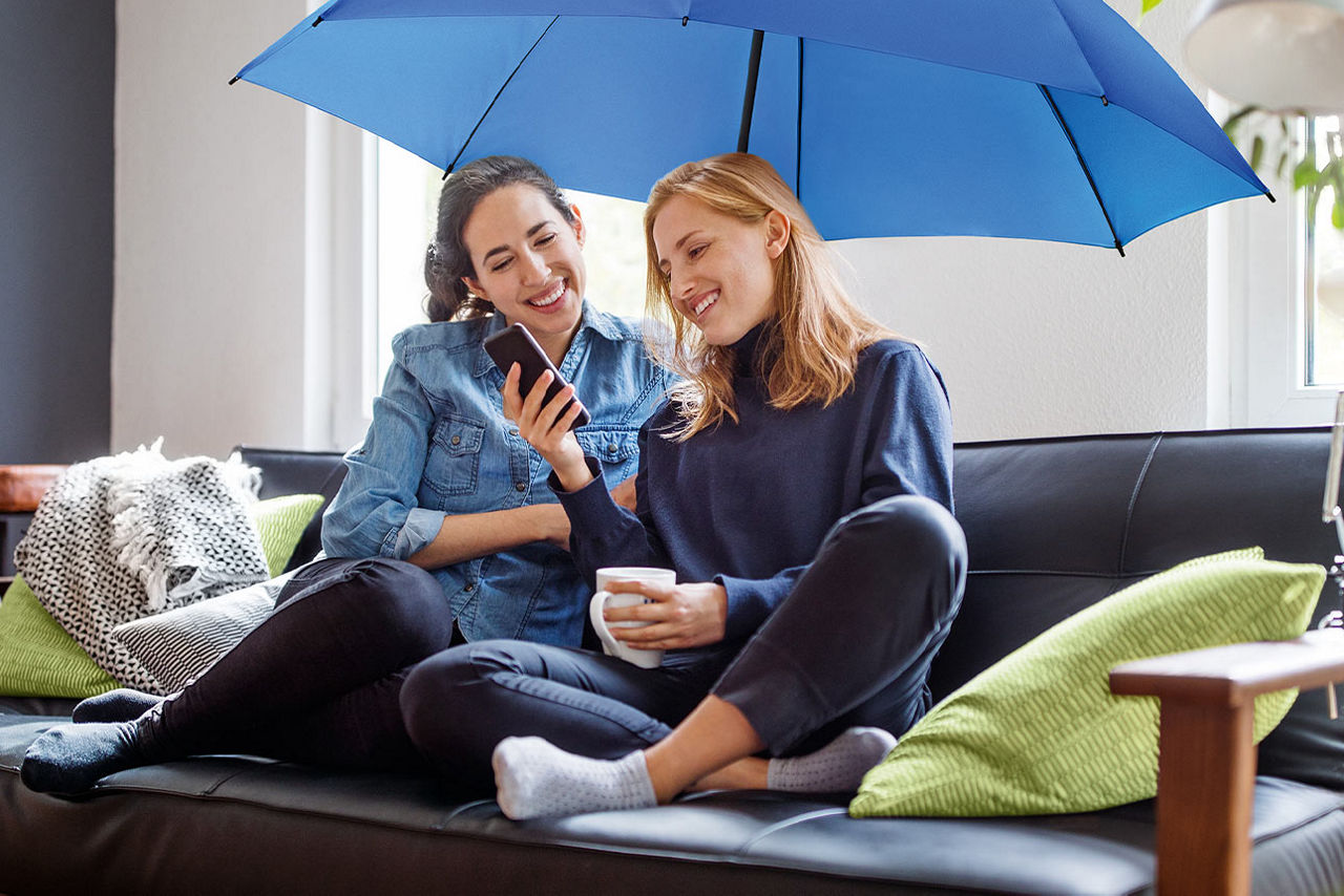 Zwei Frauen sitzen unter einem blauen Schirm auf dem Sofa und schauen lächelnd aufs Smartphone
