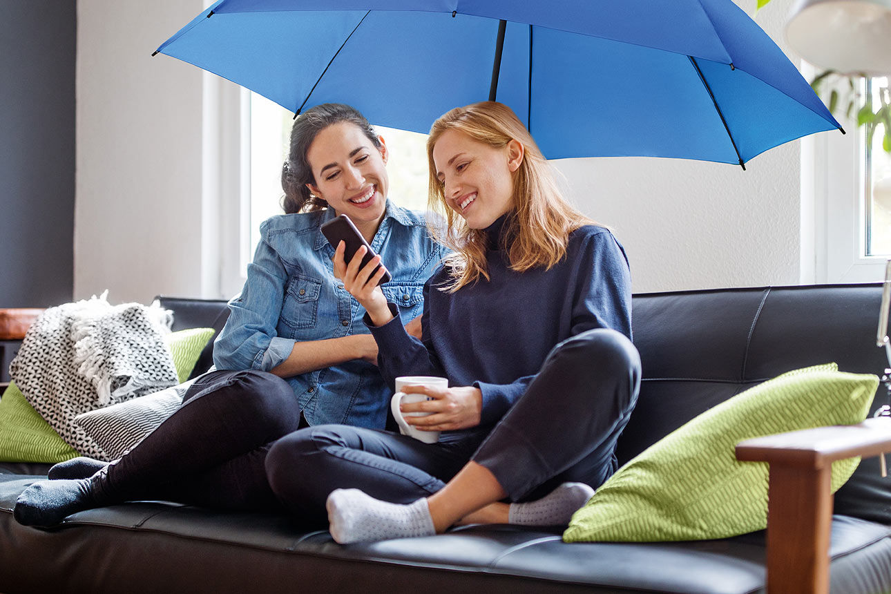 Zwei Frauen sitzen unter einem blauen Schirm auf dem Sofa und schauen lächelnd aufs Smartphone