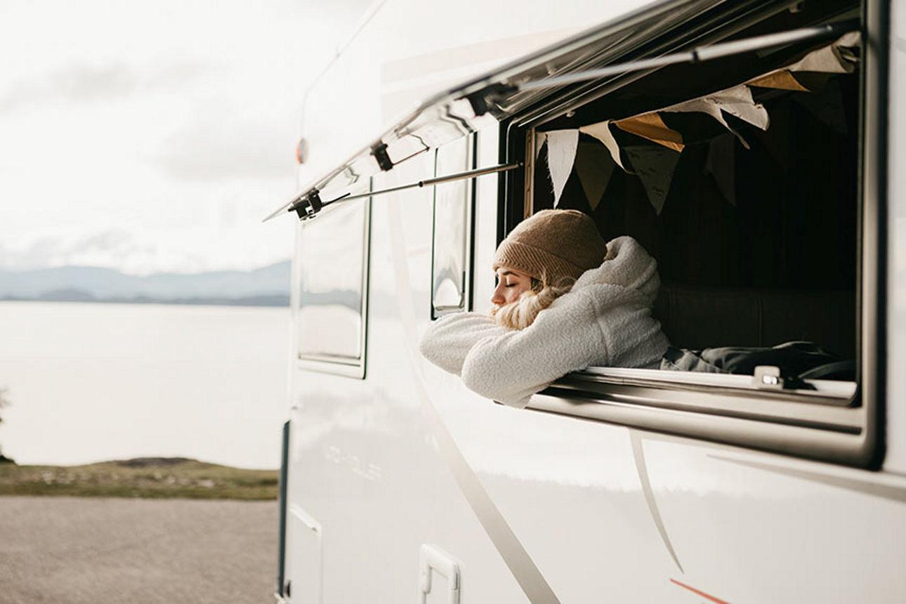 Junge Frau lehnt mit verschränkten Armen aus dem geöffneten Fenster eines Wohnwagens