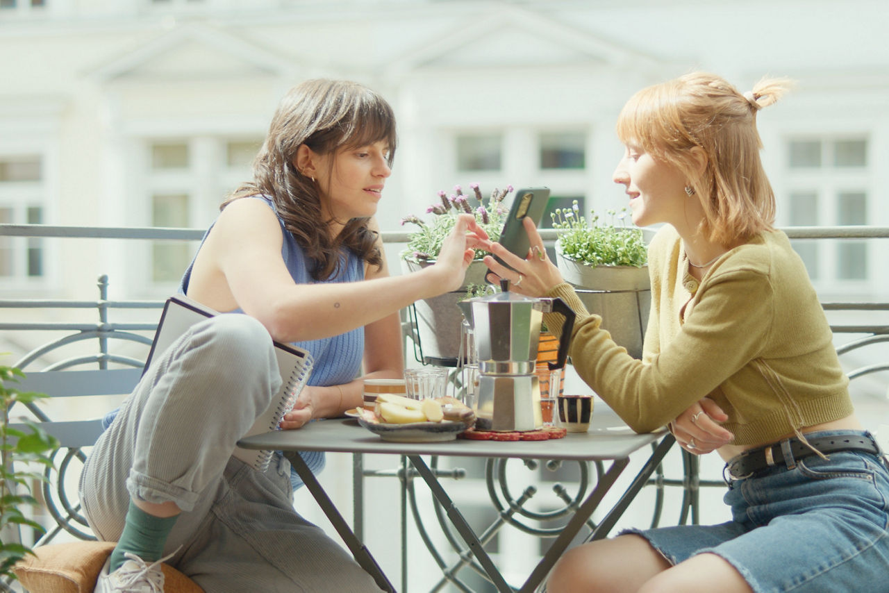 Zwei junge Frauen sitzen sich auf einem Balkon an einem mit Snacks und Kaffee gedecktem Tisch gegenüber. Die Rechte zeigt der Linken etwas auf ihrem Handy. 