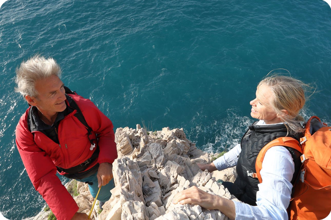 Zwei Personen stehen auf felsiger Klippe am Meer und unterhalten sich.