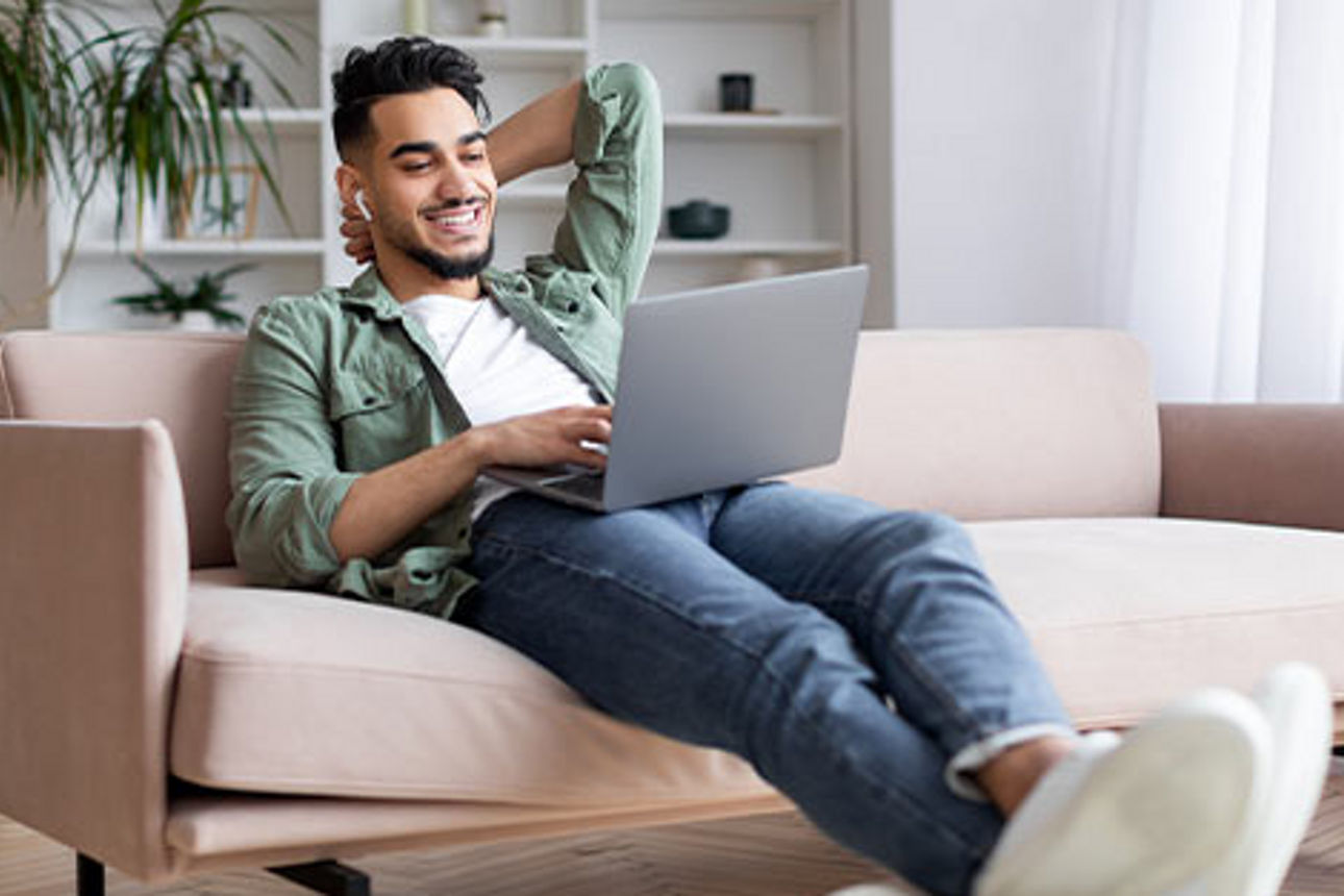 Junger Mann auf einem Sofa mit Laptop auf dem Schoß eröffnet sein Konto online