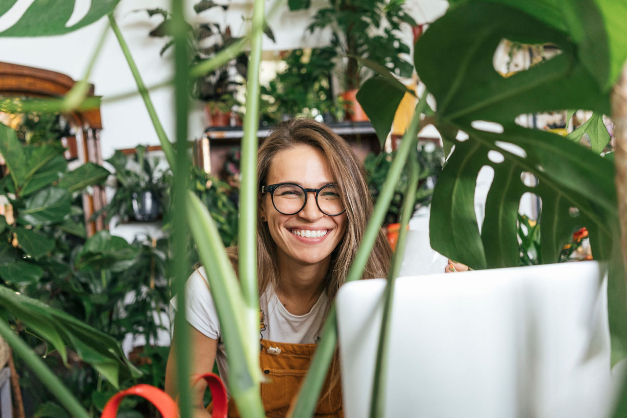 Eine Frau mit Brille blickt durch einen Vorhang aus Pflanzen auf ihr Laptop. 