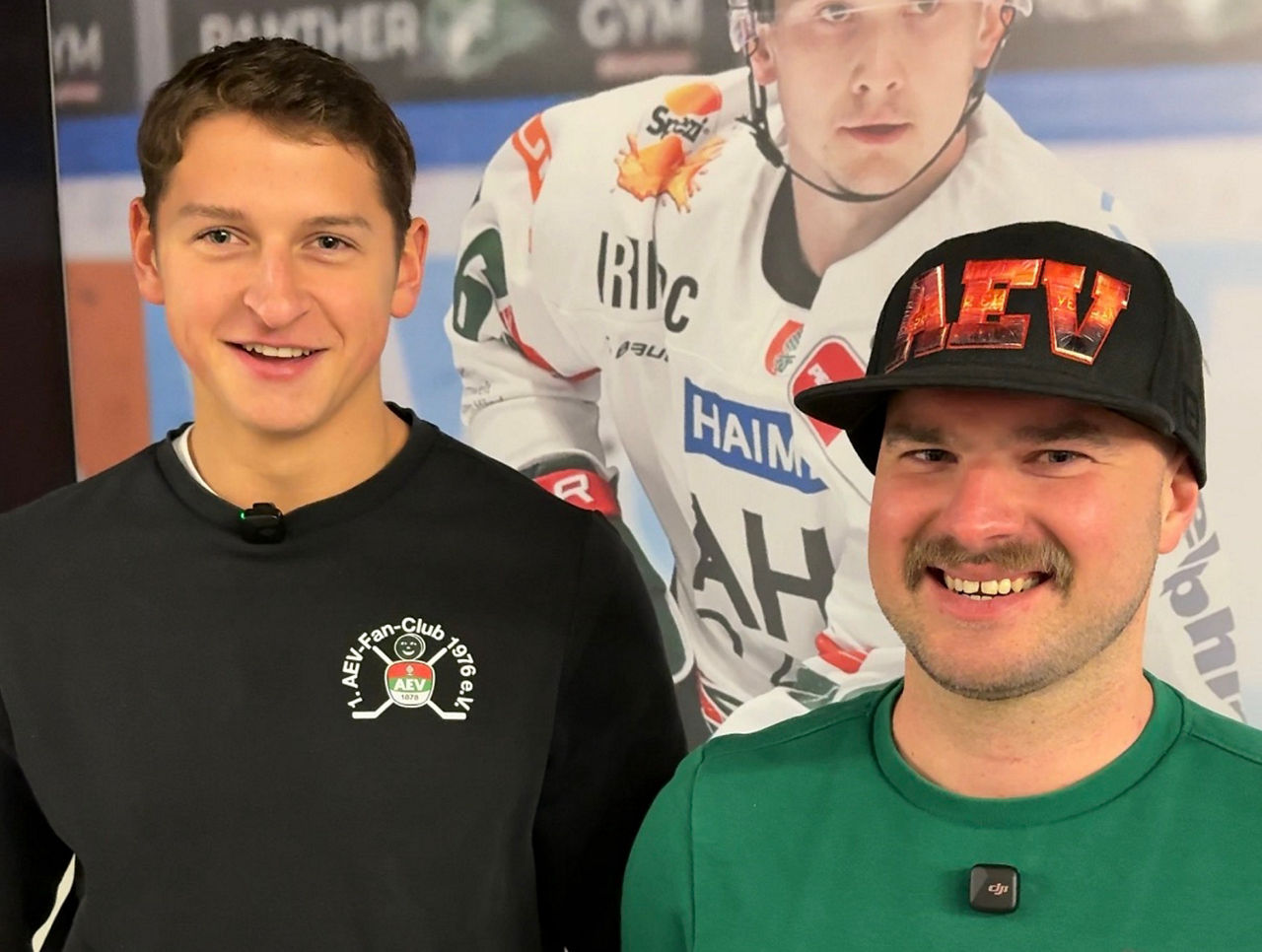 Im Hintergrund ist eine Fotowand mit einem Eishockeyspielerbild zu sehen. Im Vordergrund stehen zwei Männer, einer im schwarzen, einer im grünen Pulli.