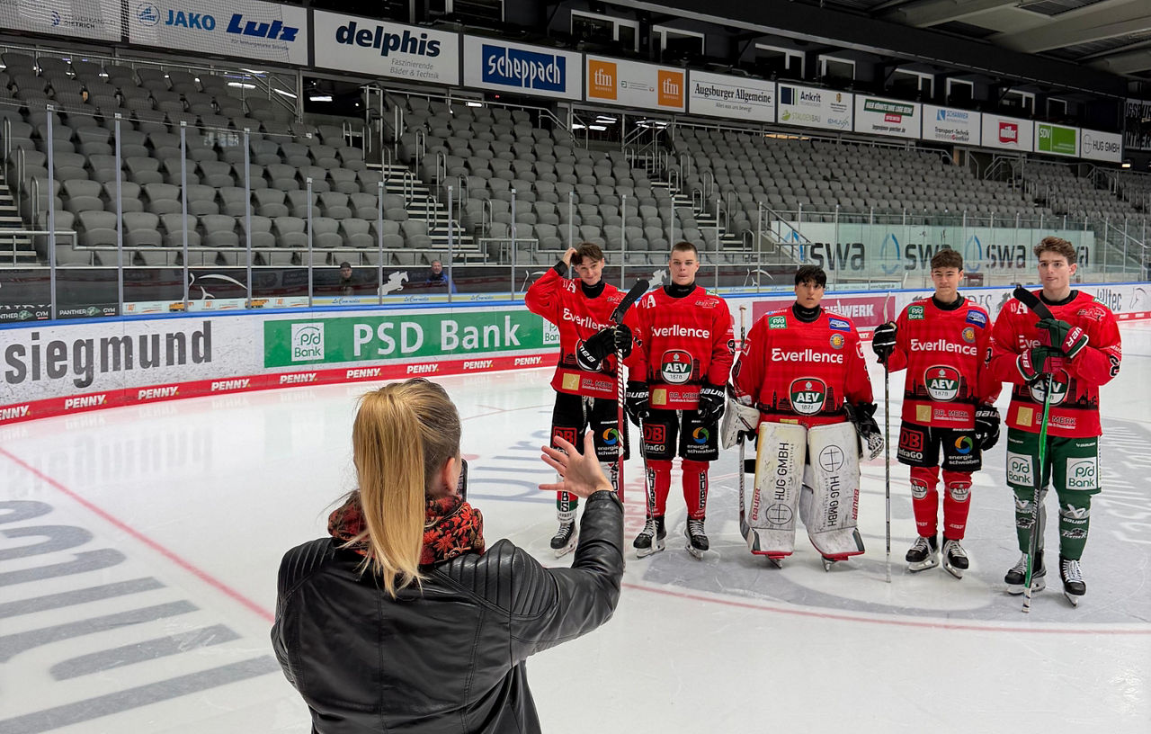 Auf dem Eis in einem Eisstadion stehen im Hintergrund fünf Nachwuchsspieler des AEV in roten Trikots in einer Reihe. Sie werden von einer Frau, die mit dem Rücken zur Kamera steht, gefilmt.