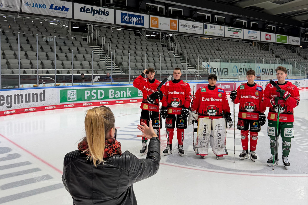 Auf dem Eis in einem Eisstadion stehen im Hintergrund fünf Nachwuchsspieler des AEV in roten Trikots in einer Reihe. Sie werden von einer Frau, die mit dem Rücken zur Kamera steht, gefilmt.