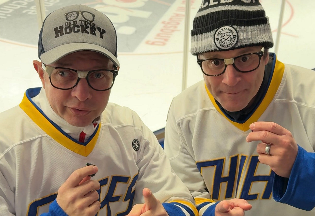 Zwei Eishockeyfans mit weißen Trikots sprechen in die Kamera. Beide halten ein kleines Mikrofon in ihren Händen. Im Hintergrund ist eine Eisfläche zu sehen.