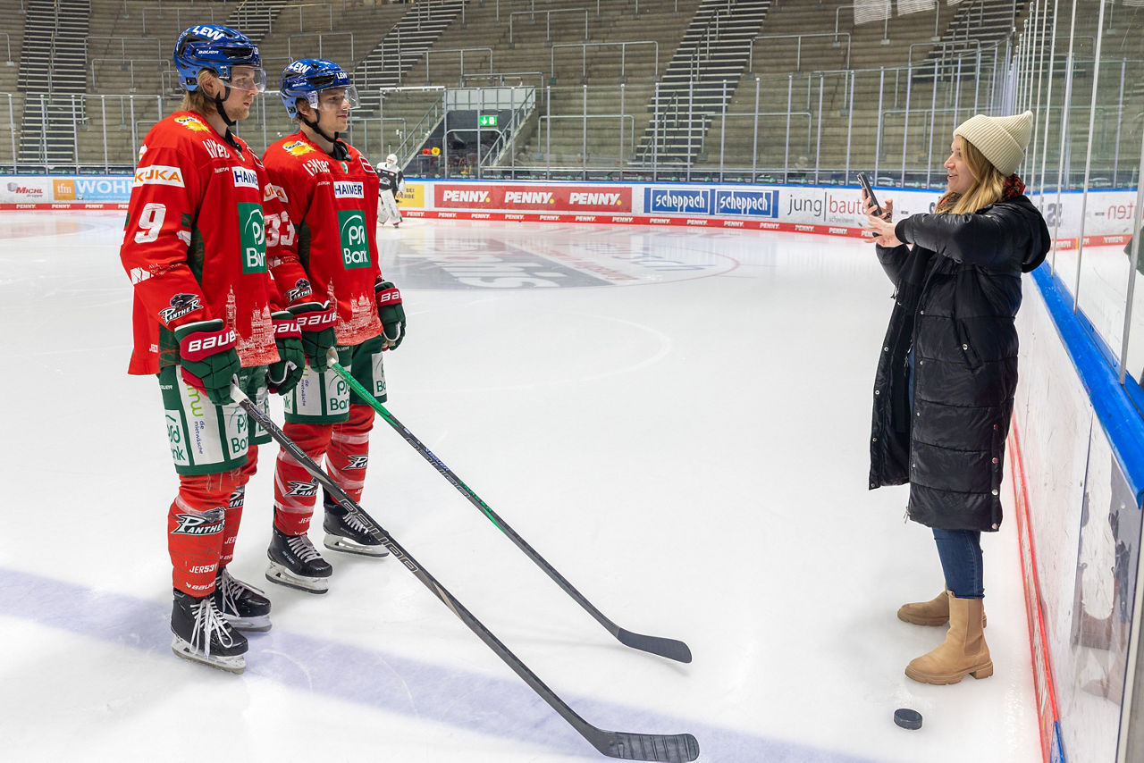 Zwei Eishockeyspieler in roten Trikots stehen in Montur auf dem Eis. Sie werden von einer blonden Frau gefilmt. Im Hintergrund ist der Stehplatzbereich zu erkennen.