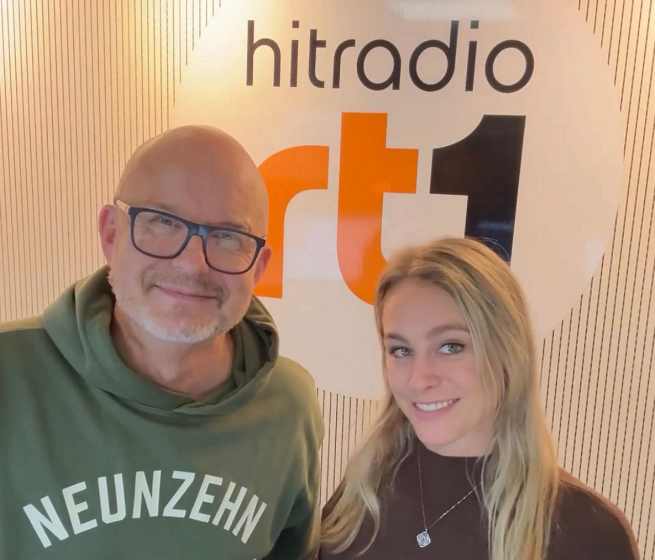 Im Hintergrund ist eine Fotowand mit einem Eishockeyspielerbild zu sehen. Im Vordergrund stehen zwei Männer, einer im schwarzen, einer im grünen Pulli.