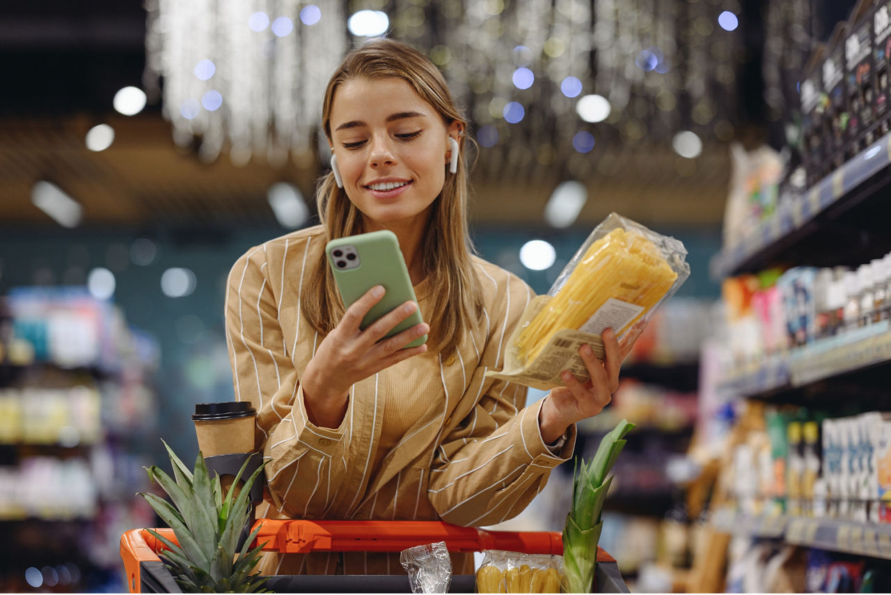 Eine junge Frau schaut im Supermarkt lächelnd auf ihr Handy, während sie einkauft.