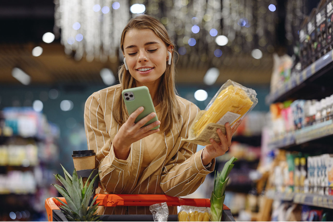 Eine junge Frau schaut im Supermarkt lächelnd auf ihr Handy, während sie einkauft.
