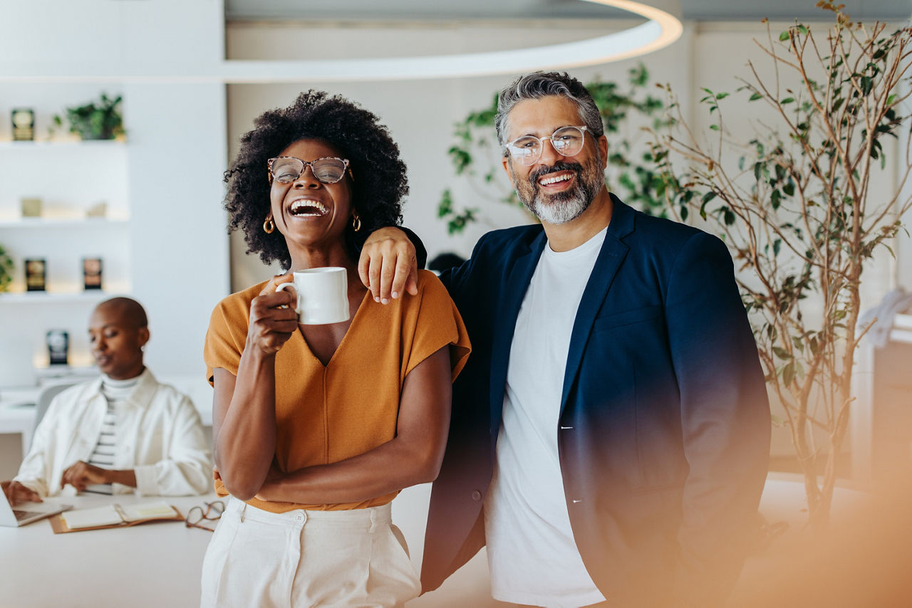 Ein Mann und eine Frau stehen nebeneinander und lachen. Die Frau hält eine Kaffeetasse in der Hand.