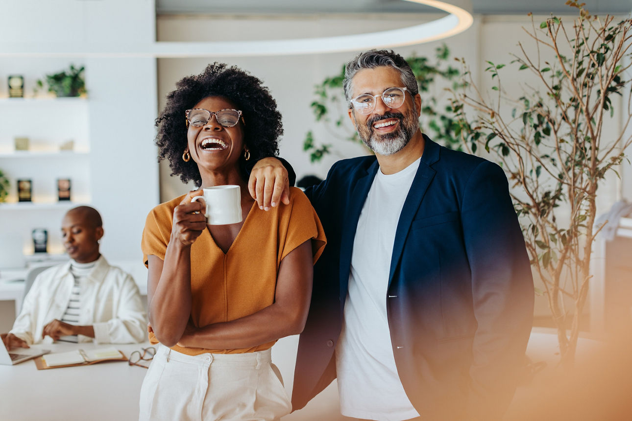 Ein Mann und eine Frau stehen nebeneinander und lachen. Die Frau hält eine Kaffeetasse in der Hand.