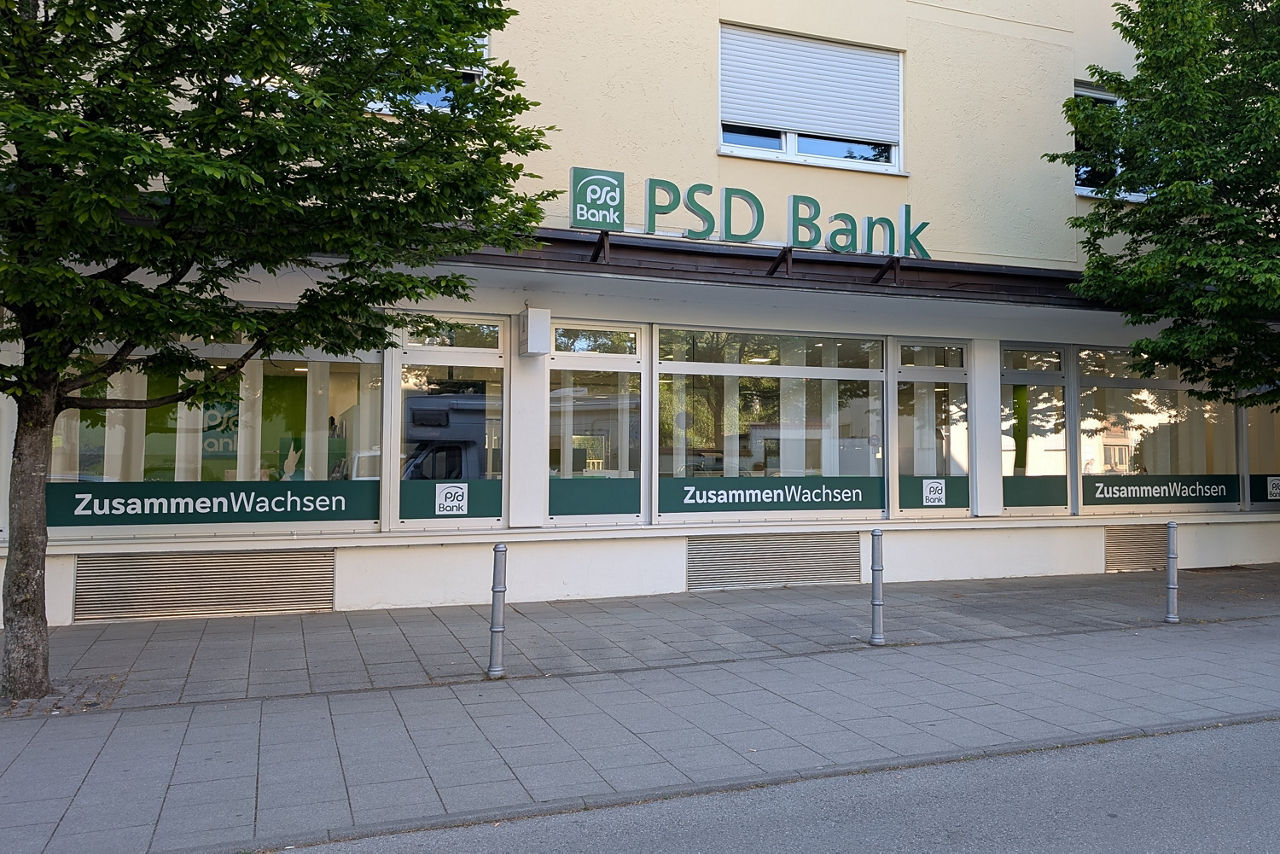 Außenfassade eines Gebäudes, in dem sich das Kundencenter der PSD Bank München befindet. Die Fenster sind unten durchgängig in grün beklebt und zeigen Abwechselnd das Word "ZusammenWachsen" sowie das Logo der PSD Bank.
