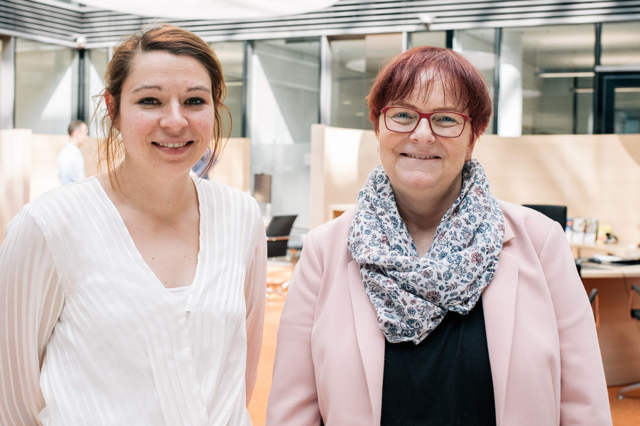Zwei lächelnde Frauen stehen in einem modernen Büro und blicken in die Kamera
