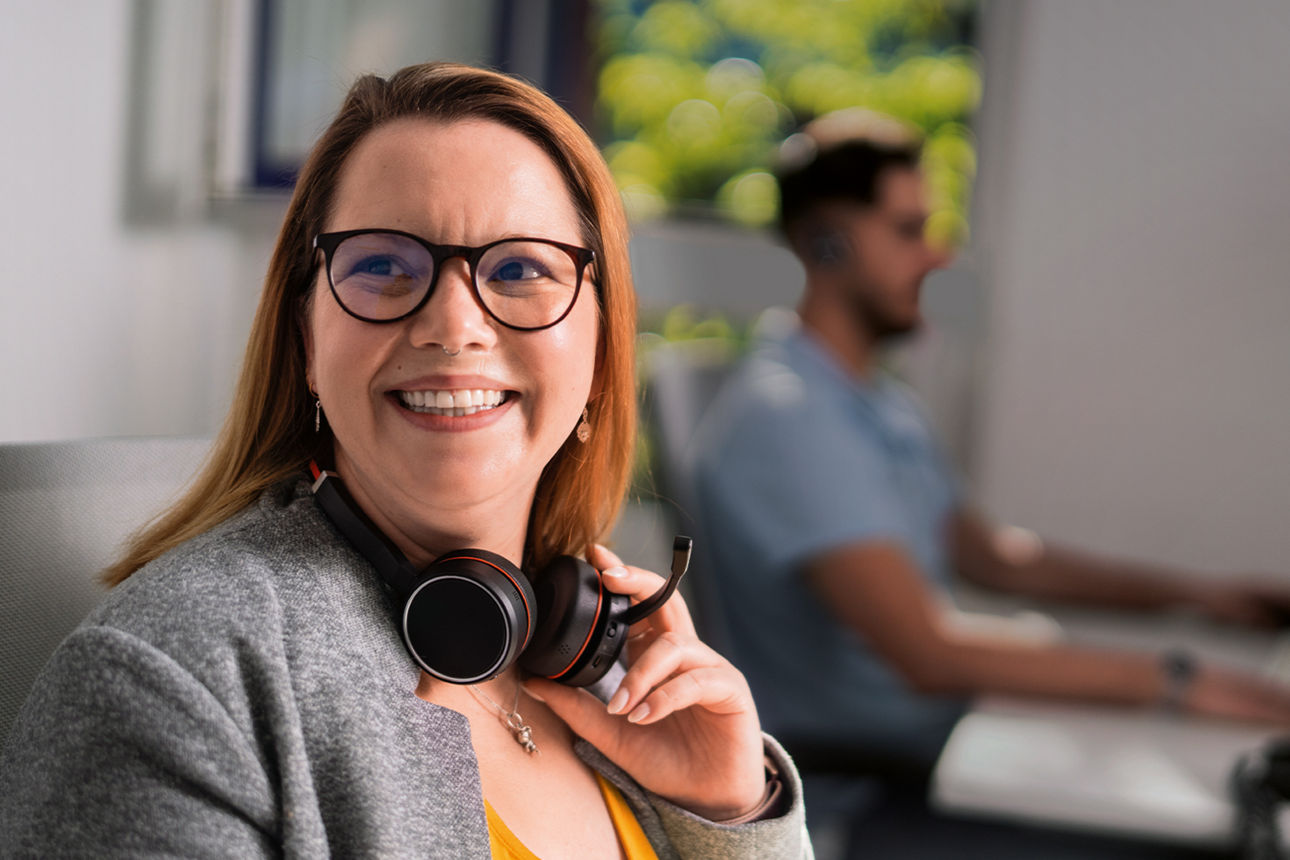 Junge Frau mit roten langen Haaren, Brille und Nasenpiercing lächelt freundlich. Eine Hand liegt auf dem Headset am Hals.