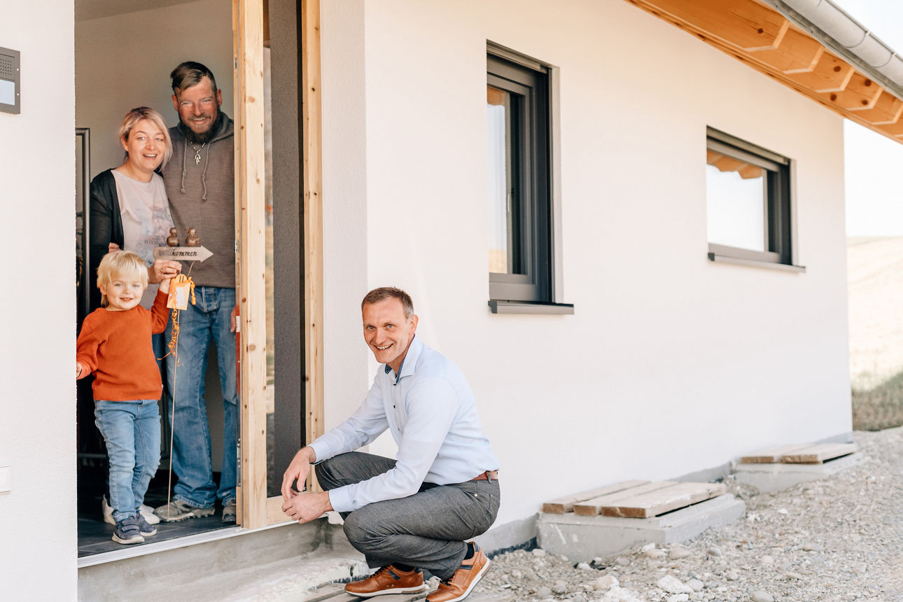 Eine junge Familie steht in der Haustür Ihres Neubaus und erhält vom Wohnbaufinanzierungsberater ein Einzugsgeschenk.