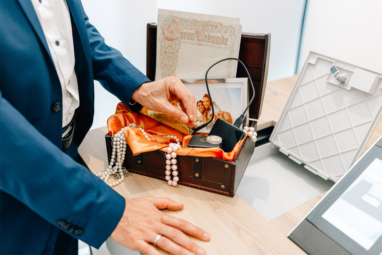 Eine Schatztruhe mit verschiedenen Wertgegenständen und Hände die etwas hineinlegen.