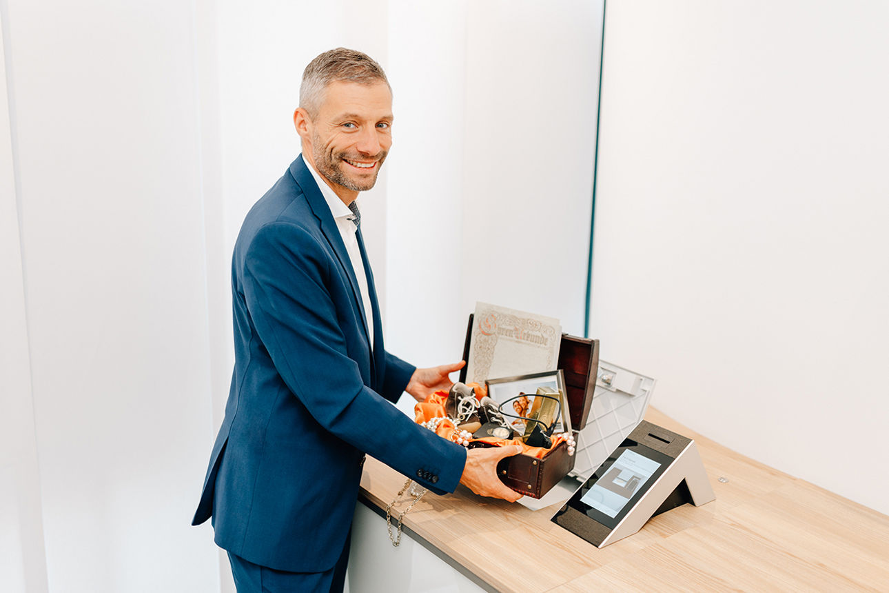 Ein lächelnder Mann hält eine Schatztruhe in der Hand mit vielen Schätzen. In einem Raum für Schließfächer der Bank