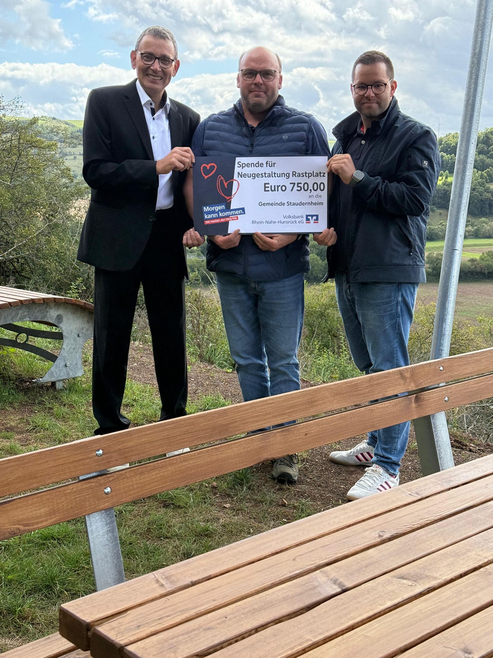 Drei Erwachsene stehen auf einer Wiese und halten einen Spendenscheck in der Hand.