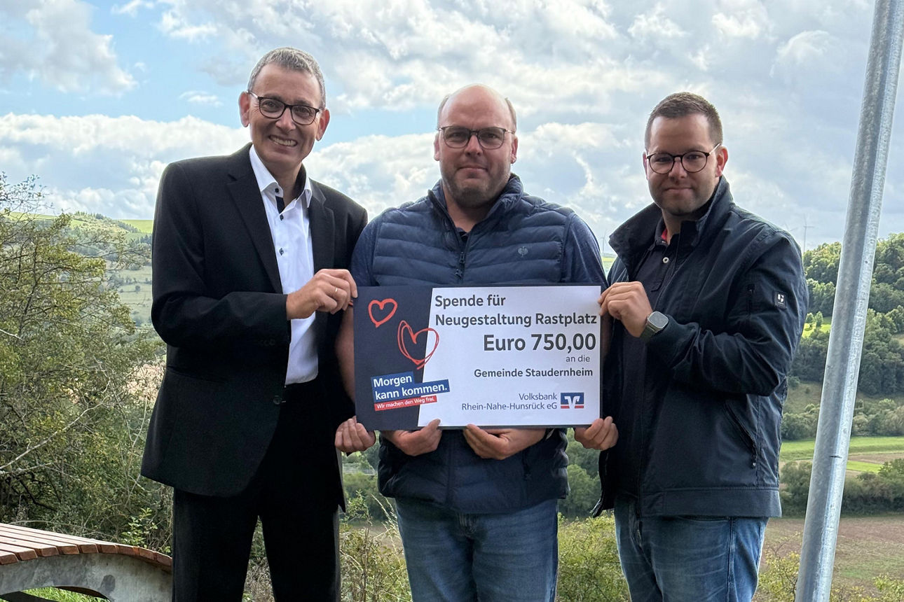 Drei Erwachsene stehen auf einer Wiese und halten einen Spendenscheck in der Hand.