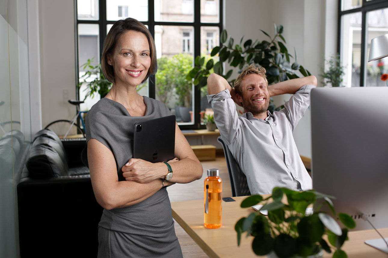 Eine Frau mit Aktentasche steht in einem Büro, hinter ihr sitzt ein Mann lächelnd an einem Schreibtisch