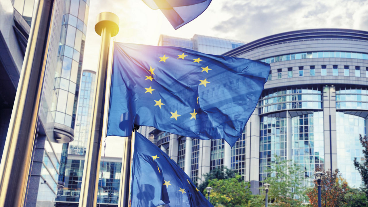 Blaue Flaggen der EU mit gelben Sternen im Kreis vor modernen Gebäuden mit großen Glasfronten