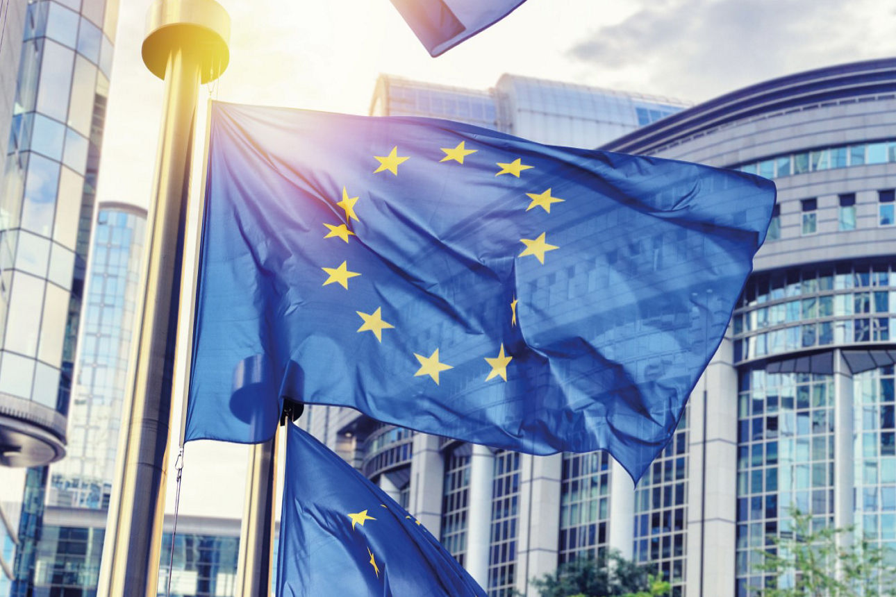 Blaue Flaggen der EU mit gelben Sternen im Kreis vor modernen Gebäuden mit großen Glasfronten