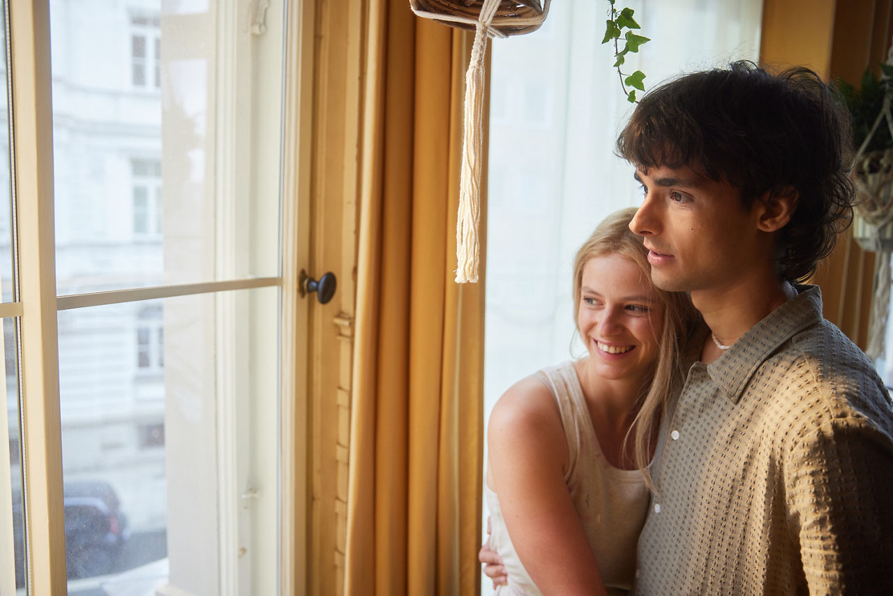 Ein Pärchen steht in einer hellen Wohnung am Fenster. Sie umarmt ihn von der Seite. Gemeinsam Blicken Sie aus dem Fenster auf die Straße. Ein Pärchen steht in einer hellen Wohnung am Fenster. Sie umarmt ihn von der Seite. Gemeinsam Blicken Sie aus dem Fenster auf die Straße.