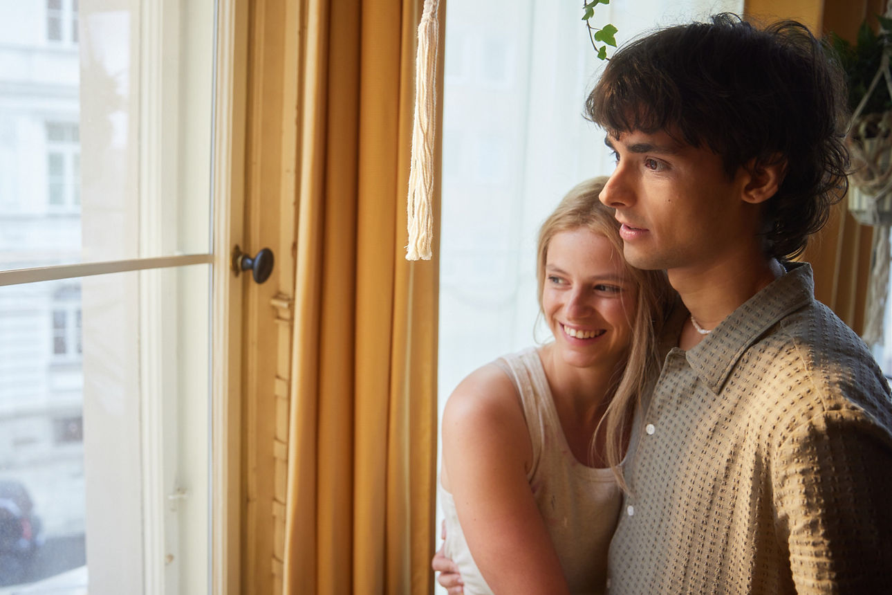 Ein Pärchen steht in einer hellen Wohnung am Fenster. Sie umarmt ihn von der Seite. Gemeinsam Blicken Sie aus dem Fenster auf die Straße. Ein Pärchen steht in einer hellen Wohnung am Fenster. Sie umarmt ihn von der Seite. Gemeinsam Blicken Sie aus dem Fenster auf die Straße.