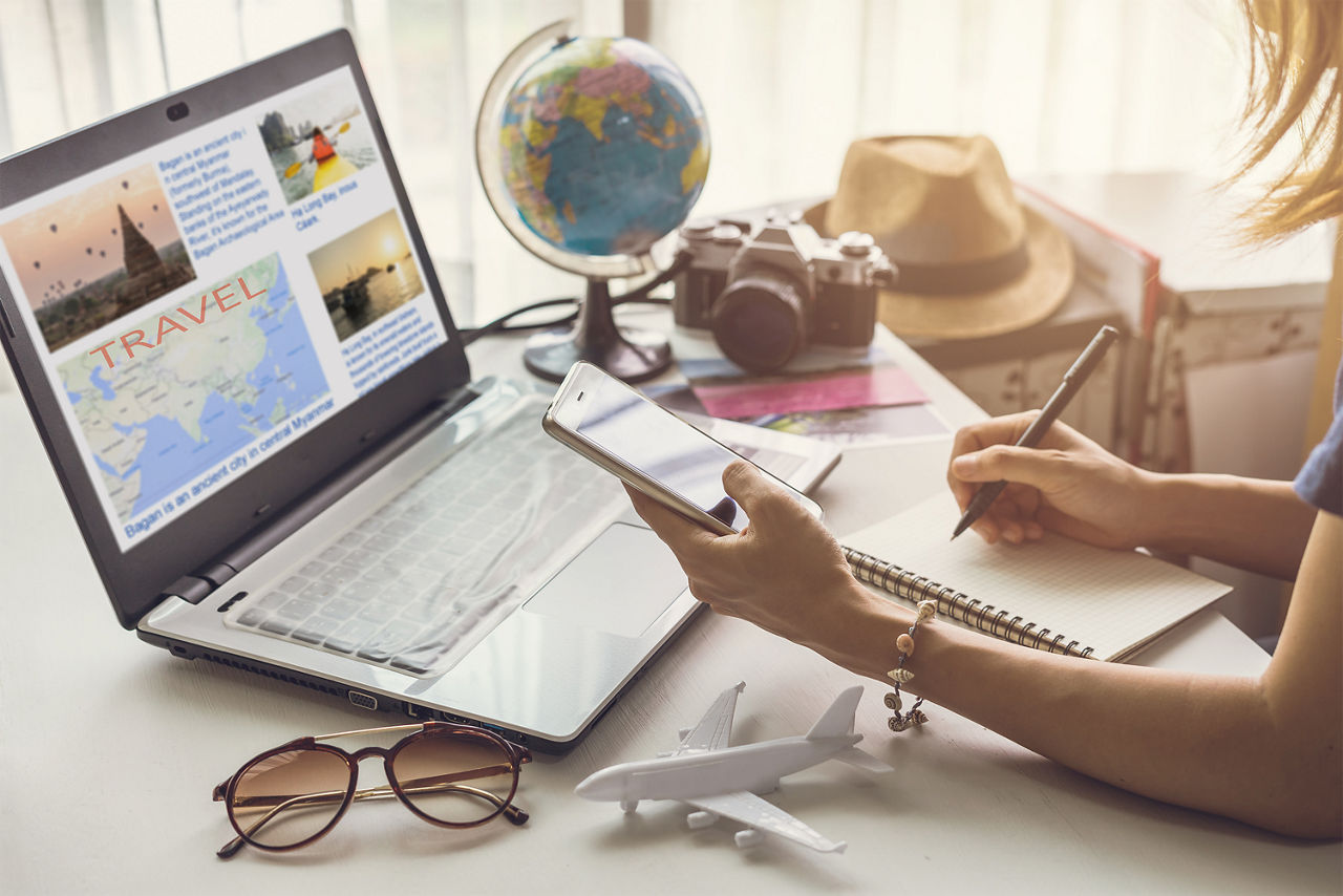 Eine junge Frau sitzt vor einem Laptop mit einem Handy in der Hand. Vor ihr liegen eine Sonnenbrille, ein Modellflugzeug und ein Schreibblock. Im Hintergrund sieht man einen Globus, eine Kamera und einen Strohhut.