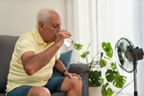 Ein älterer Mann sitzt auf einer Couch und trinkt ein Glas Wasser. Vor ihm steht ein Ventilator.