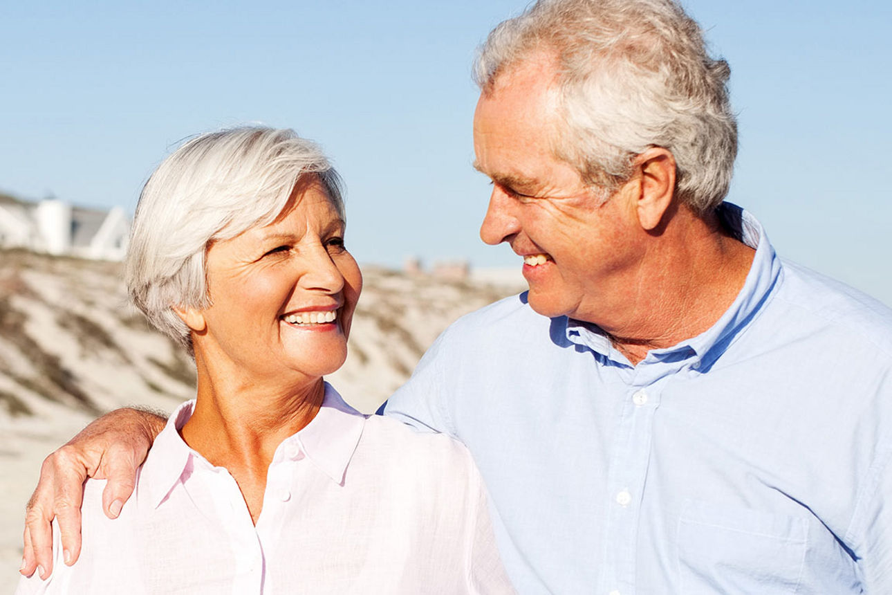 Ein älteres Paar läuft Arm in Arm über den Strand und sieht sich lächelnd an