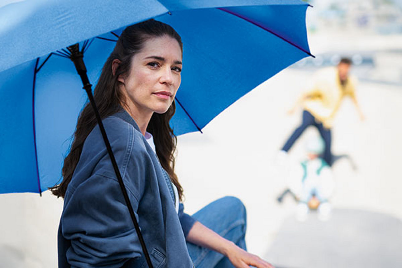 Eine junge Frau unter einem großen blauen Regenschirm, im Hintergrund ihre Familie