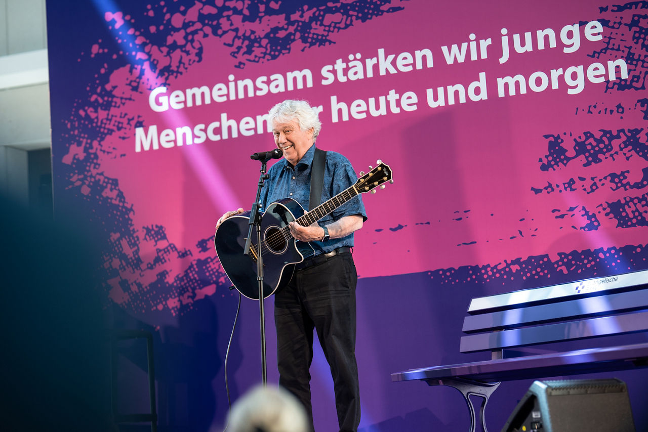 Rolf Zuckowski steht singend auf einer Bühne, hinter ihm ein großer Schriftzug