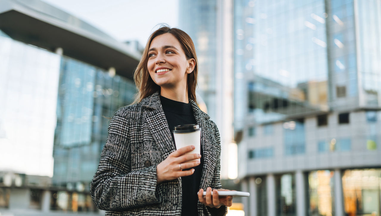 Junge lächelnde Frau mit einem Kaffeebecher in der Hand nutzt ihr Mobiltelefon im Gehen