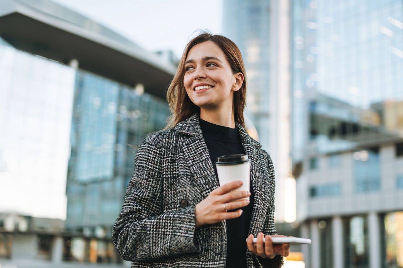 Junge lächelnde Frau mit einem Kaffeebecher in der Hand nutzt ihr Mobiltelefon im Gehen