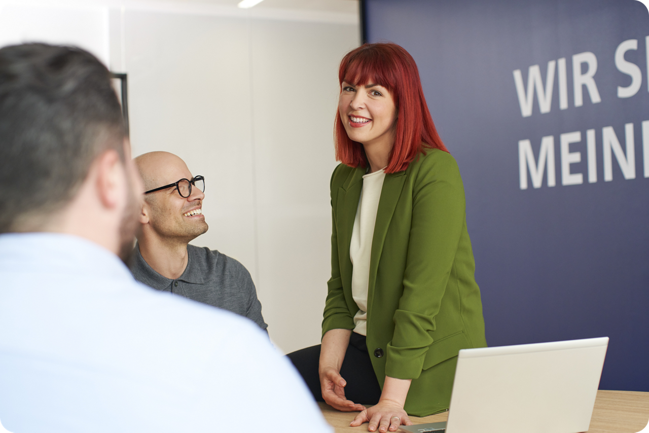 Eine junge Frau sitzt halb auf einem Tisch, zwei weitere Personen sitzen am Tisch, sie diskutieren miteinander