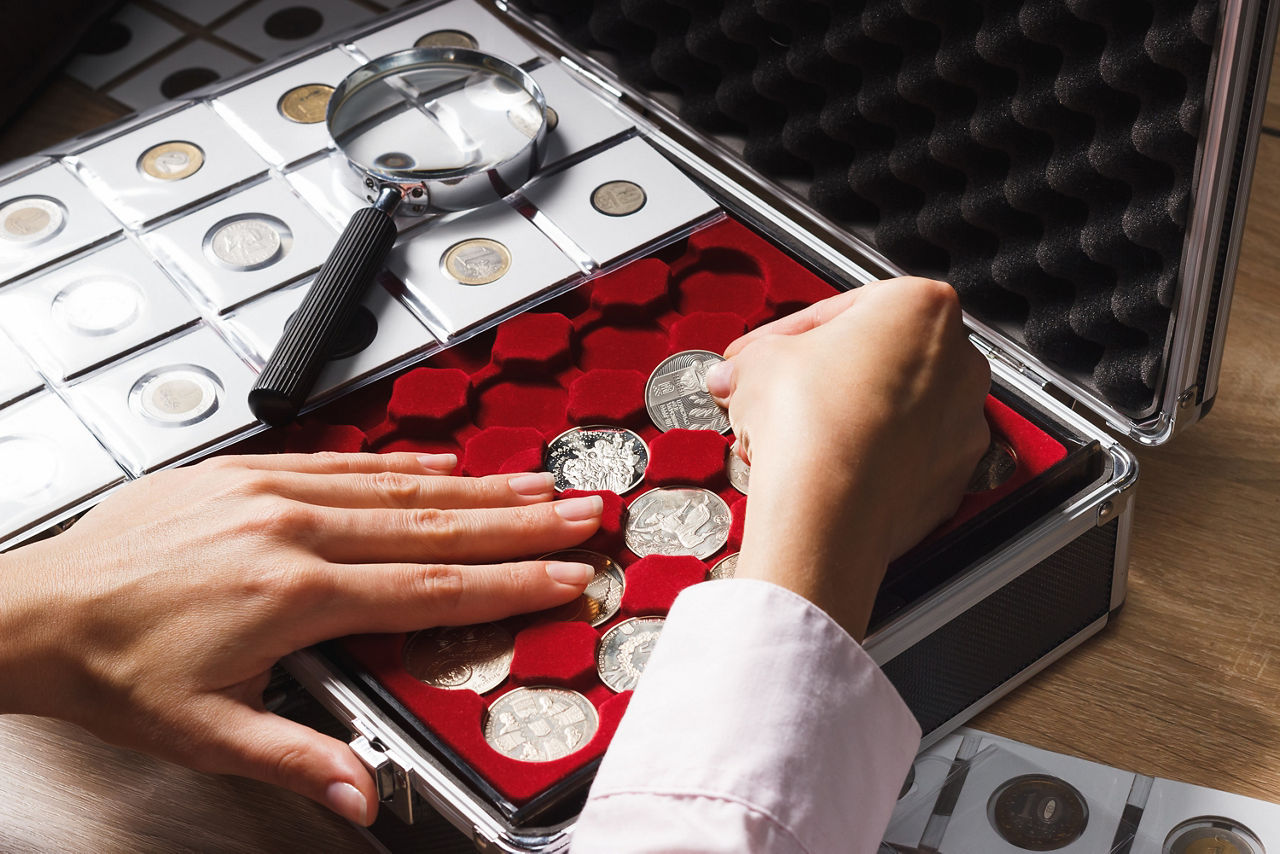 Zwei Hände sortieren Silbermünzen in eine Sammelbox mit rotem Samtfutter. Auf der Box liegt eine Lupe.