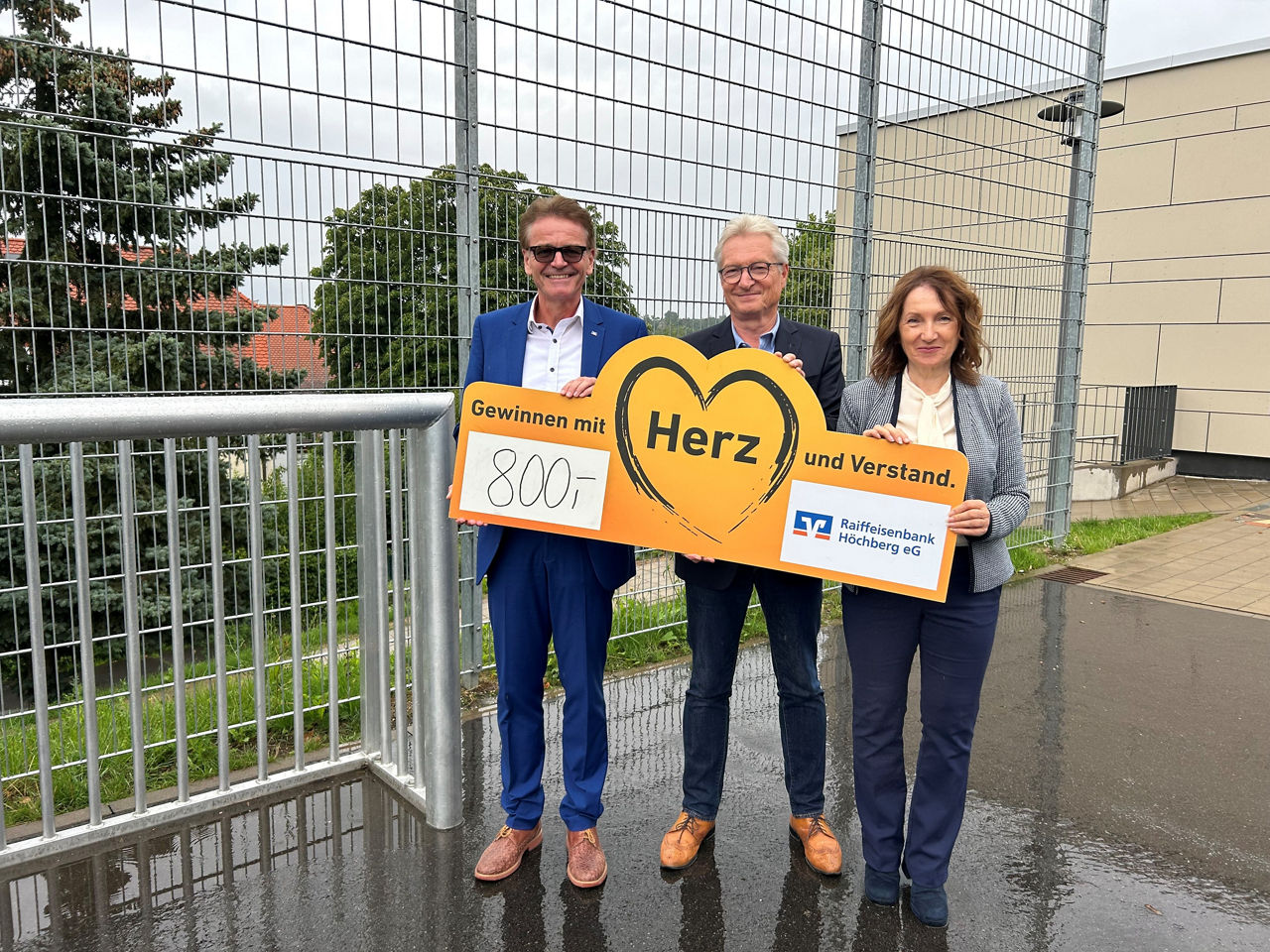 Drei Personen vor einem Tor mit einem Scheck. 