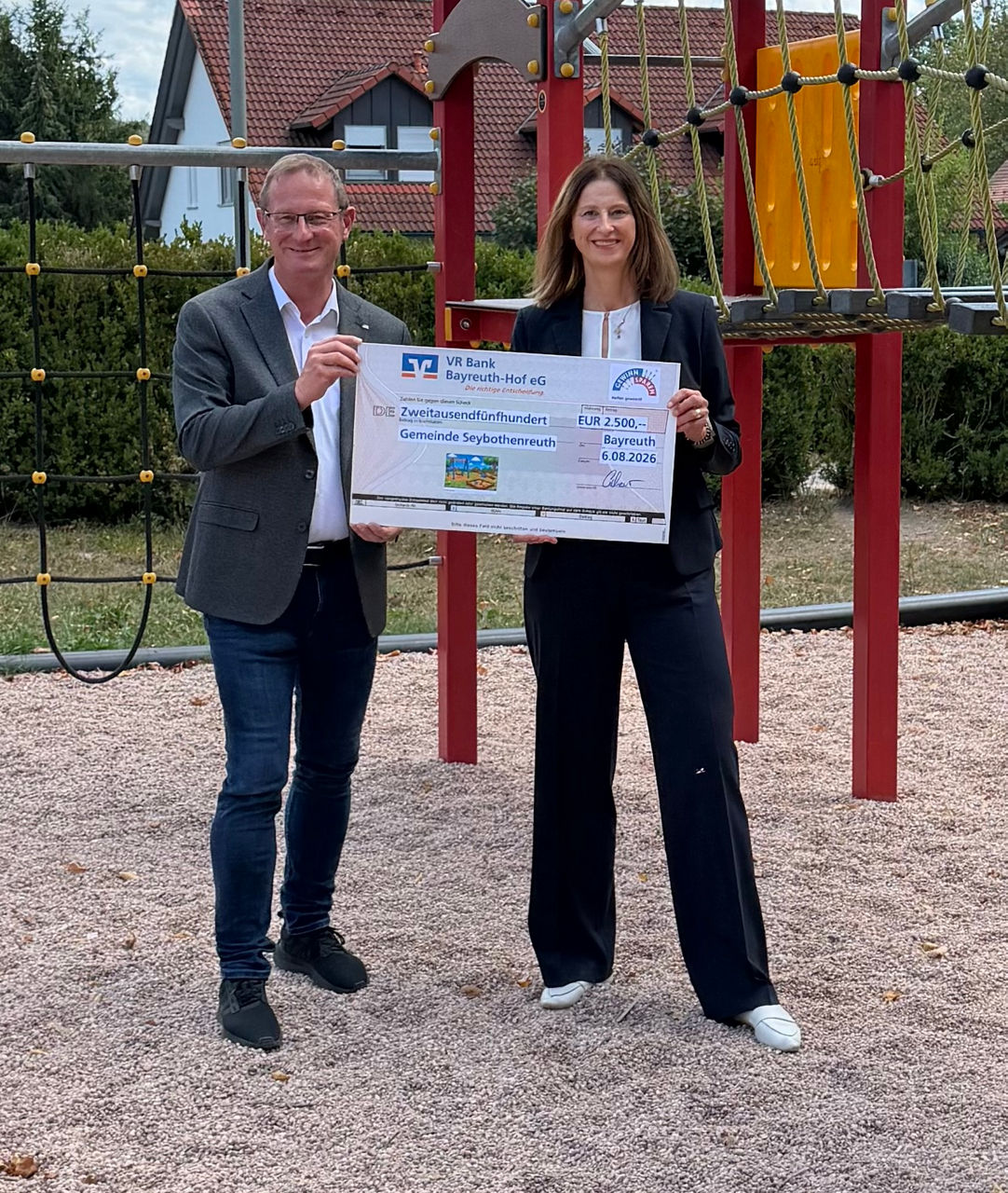 Zwei Personen stehen auf einem Spielplatz vor bunten Klettergeräten und präsentieren gemeinsam einen Spendenscheck.