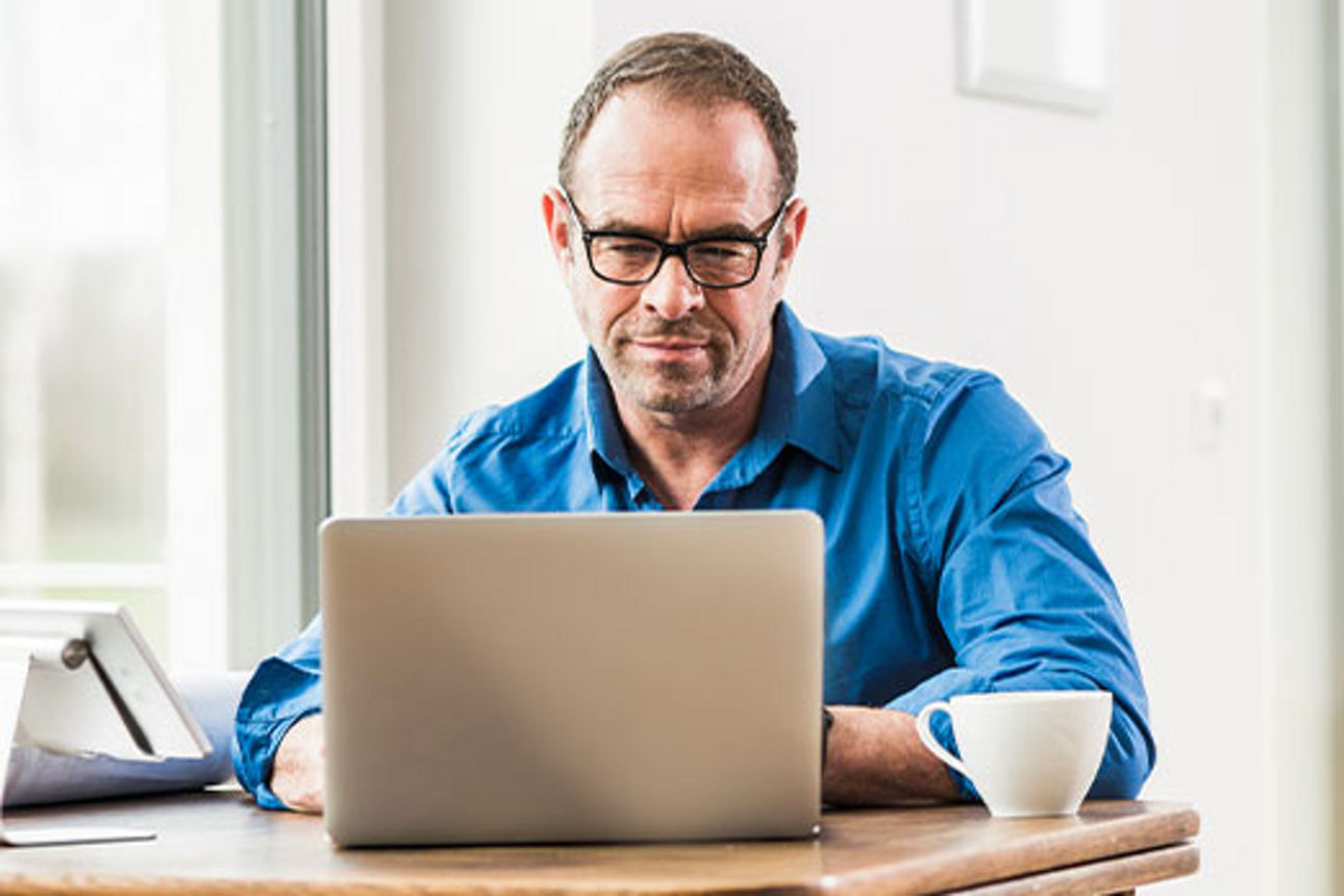 Mann sitzt mit Laptop und einer Tasse Kaffee an einem Tisch