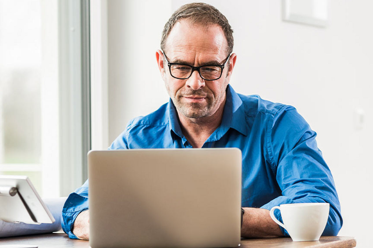 Mann sitzt mit Laptop und einer Tasse Kaffee an einem Tisch