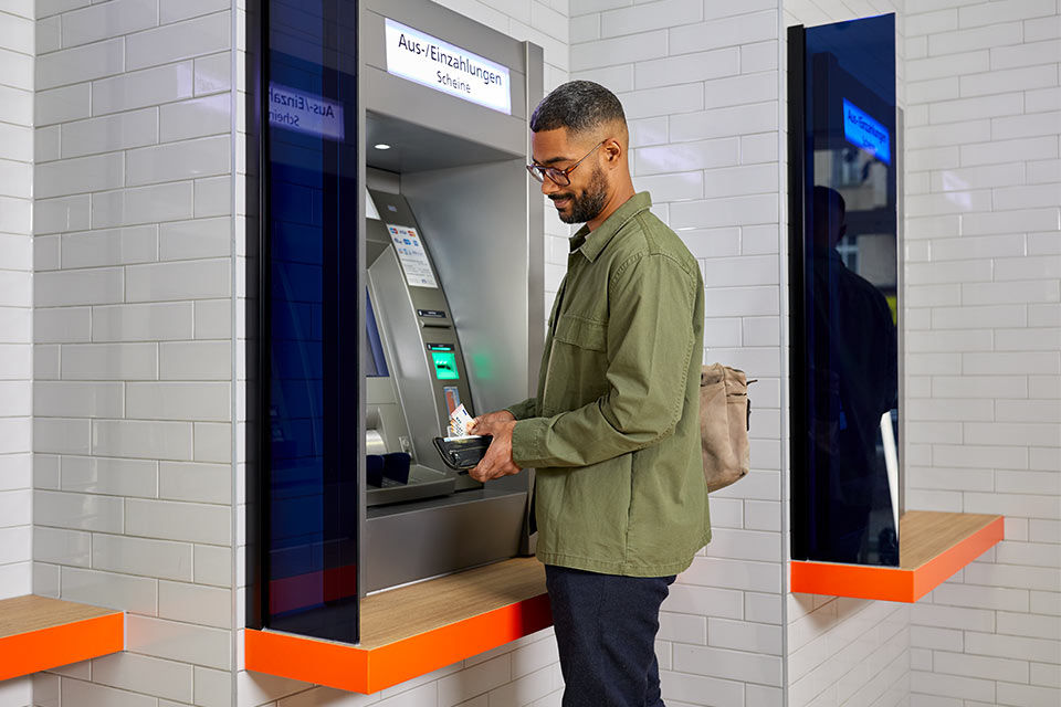 Ein junger Mann steht vor einem Geldautomaten mit einer Geldbörse und Geld in der Hand.