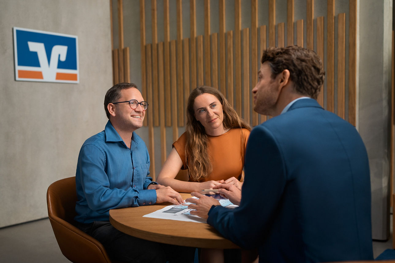 Drei Personen sitzen an einem runden Tisch in einem modernen Beratungsraum und führen ein Gespräch.